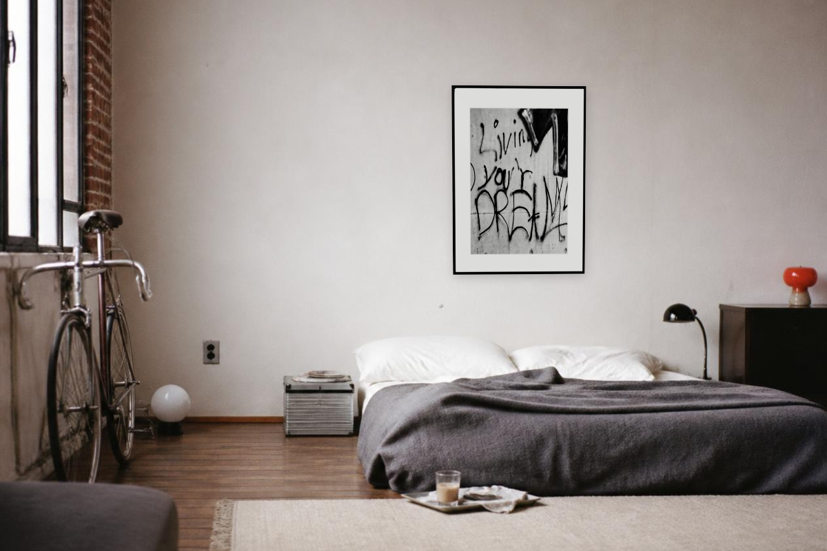 Gerahmter Fine-Art-Print mit dem Graffiti „Living your Dream“ an einer hellen Wand in einem minimalistischen Schlafzimmer.